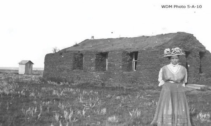 A team of two horses are pulling a plow to break Prairie topsoil. A farmer is walking behind the horses to operate the plow.