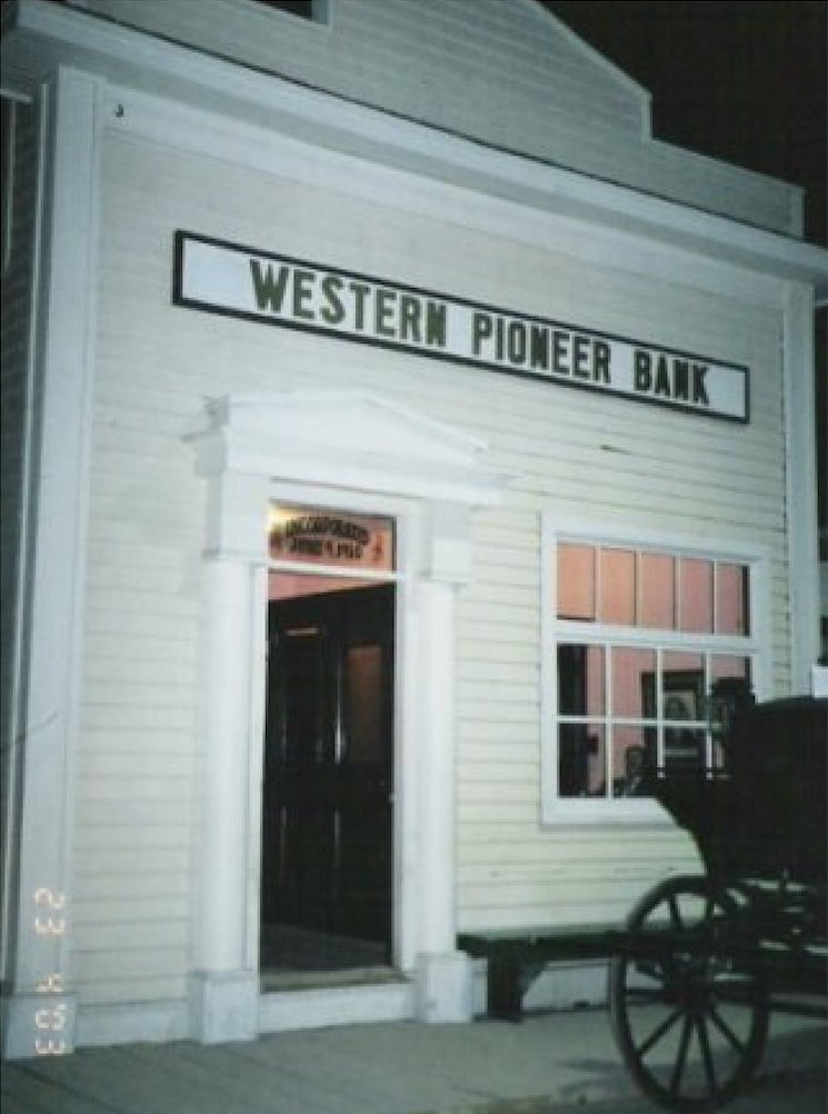 A group of men are working together to build a wooden grain elevator with piles of lumber nearby and a train in the background.