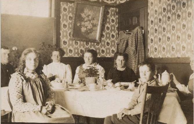 A family of eight people are sitting around a dining table for a meal together. The table is set with dishes and tea.