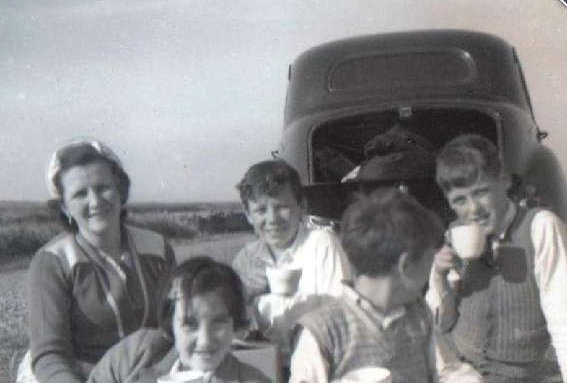 A mother and her three children are sitting down by their car to have supper in the field during harvest time.