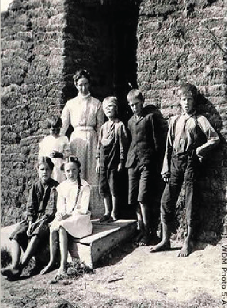 A mother and her six young children are standing in front of a sod house for a photo.