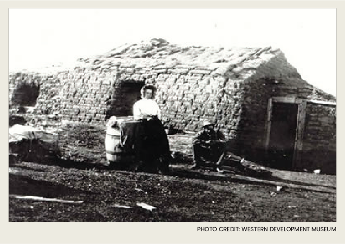 A woman and a man are standing in front of their sod house on their homestead.