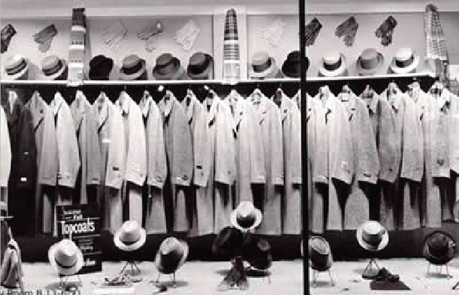 The front of a suit store is shown. The front window is large and has a big display of suits hung from a hanging rod. There are also men's accessories on display such as scarfs, hats, briefcases, and gloves.