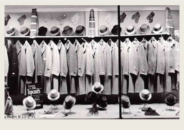 The front of a suit store is shown. The front window is large and has a big display of suits hung from a hanging rod. There are also men's accessories on display such as scarfs, hats, briefcases, and gloves.