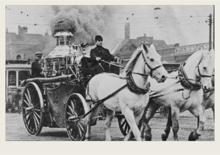 Two horses are hooked up to a steam fire engine and they are on the way to a fire