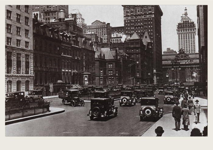 The image shows a 1920s city with a bunch of tall, brick city buildings. There are cars and people bustling around.