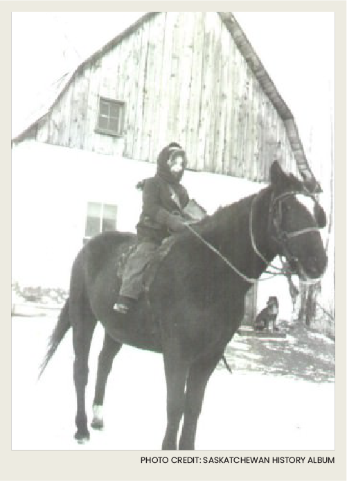 In this wintery image, a young childen is wrapped up warm in winter clothes, while sitting on a horse in front of a wooden barn.