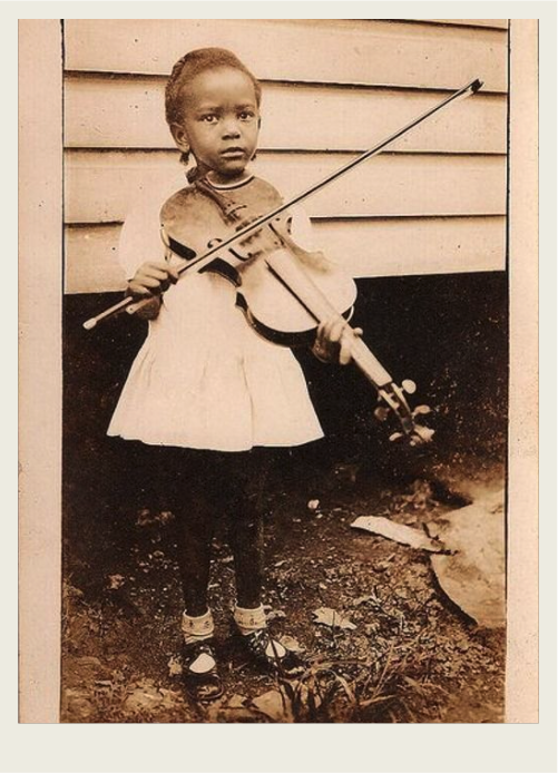 A young girl is standing by a house playing a fiddle. 