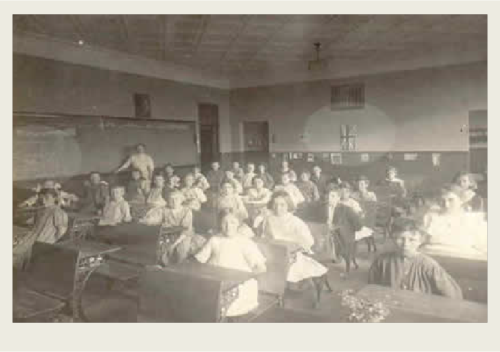 A group of about 33 childen are sitting inside a one-room school house in school desks. A teacher is standing in the background. 