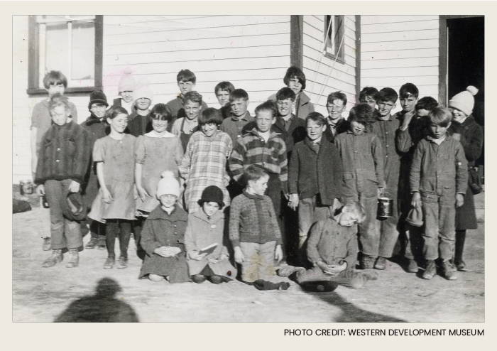 A groupd of twenty seven students are posing for a photo in front of a wooden one-room school house.