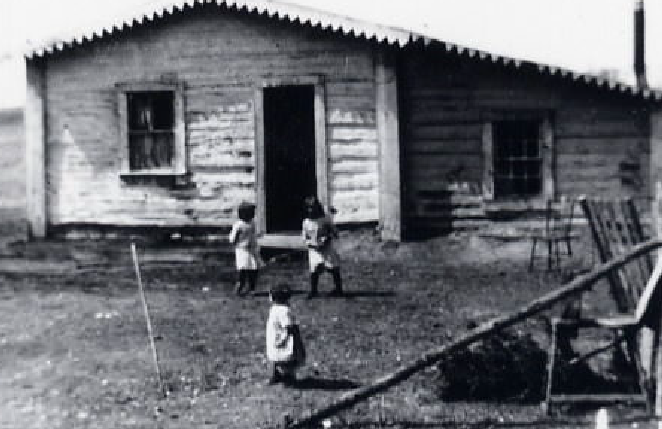 A log house on a hometead has three childen playing in front.