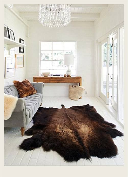 A well-lit living room with white walls and a bison hide as the rug, which is the focal point of the room. There is a small wooden shelf and a grey couch along the walls as well as a chandelier on the ceiling.