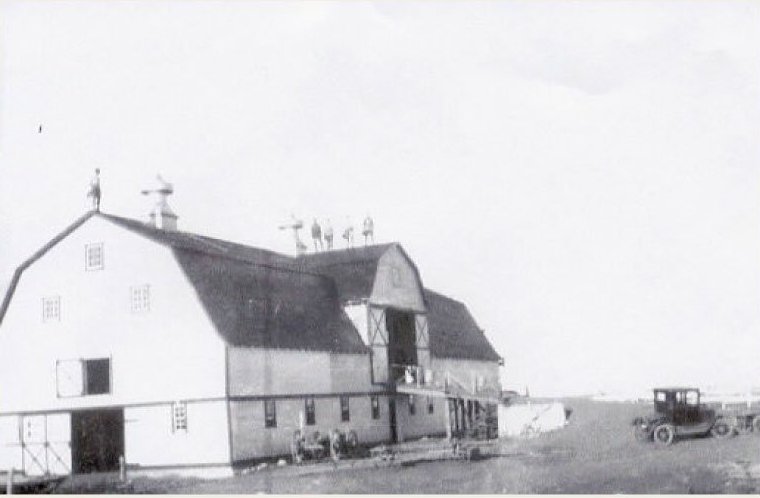 The front of a suit store is shown. The front window is large and has a big display of suits hung from a hanging rod. There are also men's accessories on display such as scarfs, hats, briefcases, and gloves.