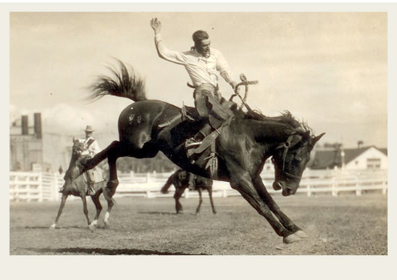 Past-Ranching-Rodeos-Overview A cowboy rides a bucking horse with one hand in the air and the other hand on the saddle. There is a white fence and a cowboy on a horse in the background.