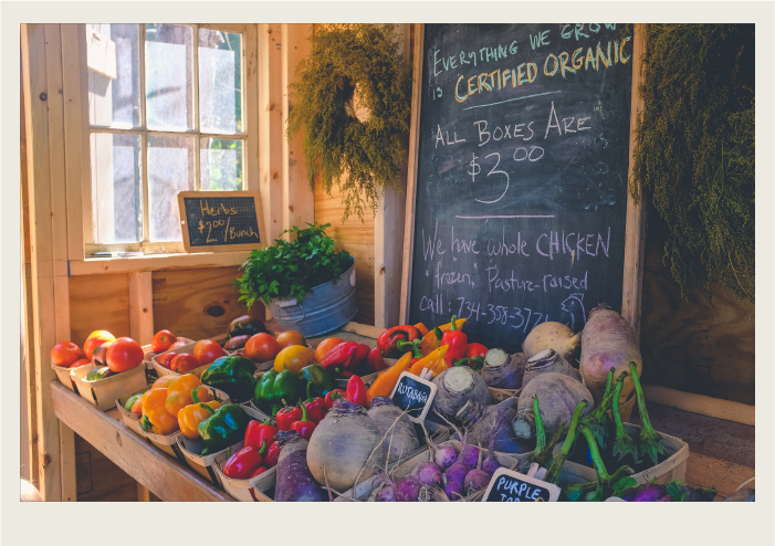 A variety of vegetables including peppers, tomatoes, and radishes are being sold at a farmers’ market with a chalkboard sign in the background with notes on it about what the farmer sells.