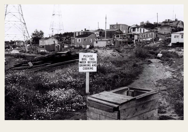 Past-Immigration-AfricanCanadians-History-community A number of small houses are shown on a hillside. There’s a wooden well with a sign next to it that reads, ‘Please boil this water before drinking and cooking’.