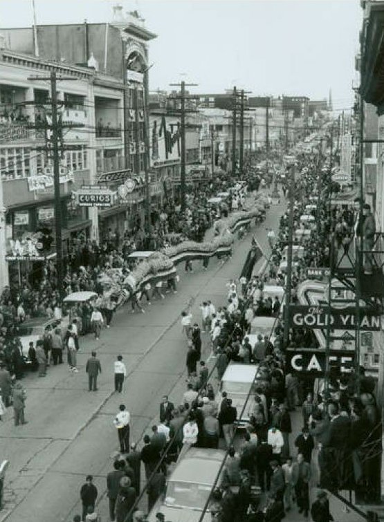 An aerial view of a parade in Chinatown in the 1920s shows many Chinese people gathered on the sides of the road as a giant dancing Dragon costume walks up the road.