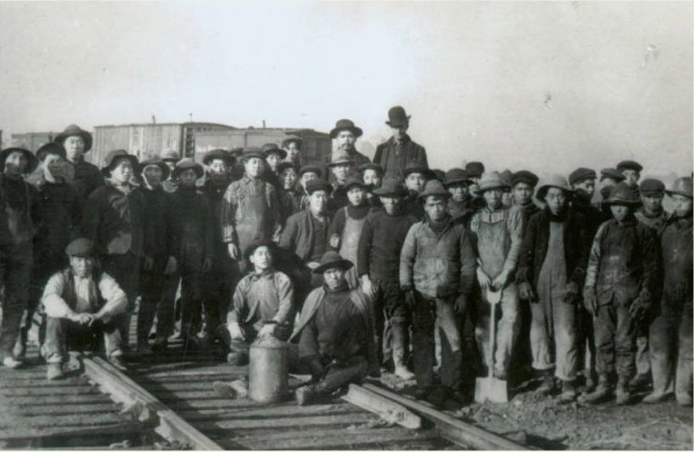 A group of many Chinese men are standing together on the railway to pose for a photo together. Most wear dirty coveralls and wide-brimmed hats.