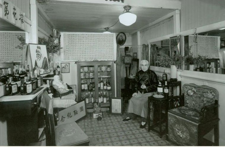 A Chinese man sits inside his small store next to cabinets that have goods and bottles in them that are for sale.