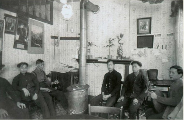 A group of nine Chinese men sit on chairs inside a home while they visit.