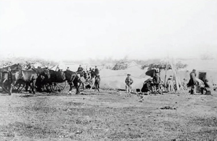 A group of men can be seen working on the prairie with oxen, horses, wagons, and a building in the background.