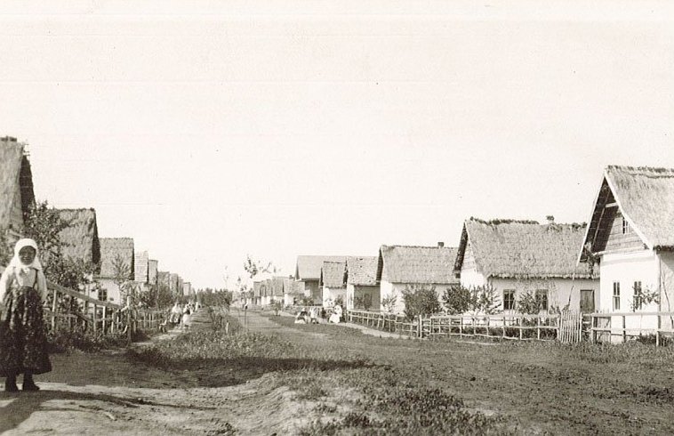 Two rows of small wooden houses are shown with a young Doukhobor girl standing along a path that goes between the houses.