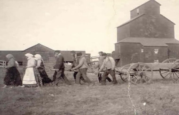 A group of four or so women and six or so men are pulling a wooden wagon together. There is a wooden grain elevator in the background.