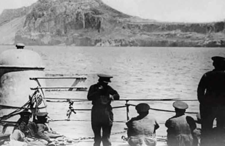 A group of four men look over the railing on a ship. The men are looking towards the coastline, which can be seen in the distance.