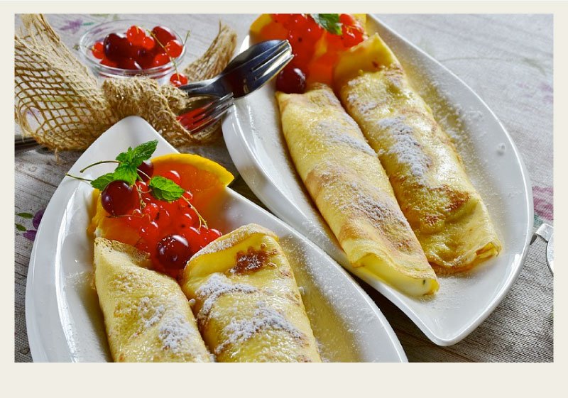 Two white dishes with two crepes each sit on a wooden table. The crepes are lightly dusted with powdered sugar.