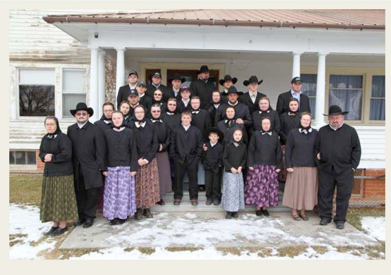 Past-Immigration-Hutterite-history-group A group of women, men, and children are grouped in front of a Hutterite colony wearing clothing that meets the clothing codes in these colonies