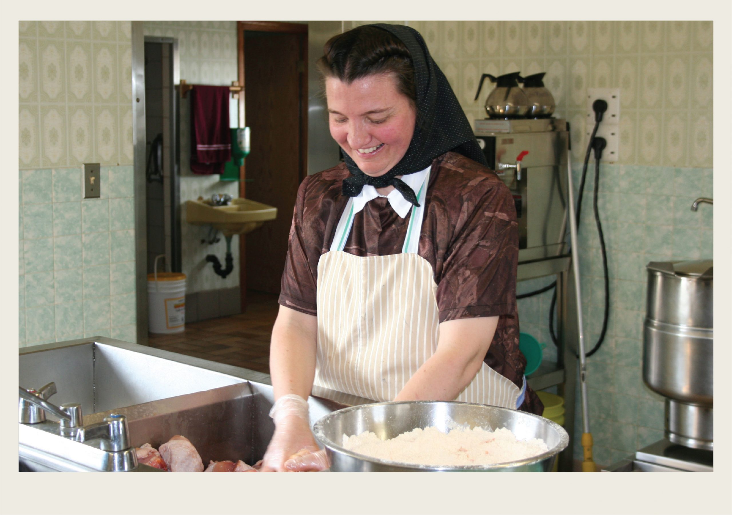 Past-Immigration-Hutterites-Settlement (HeartofSK) A Hutterite woman is in a kitchen preparing a sink full of chicken pieces, with a bowl full of flour next to her.