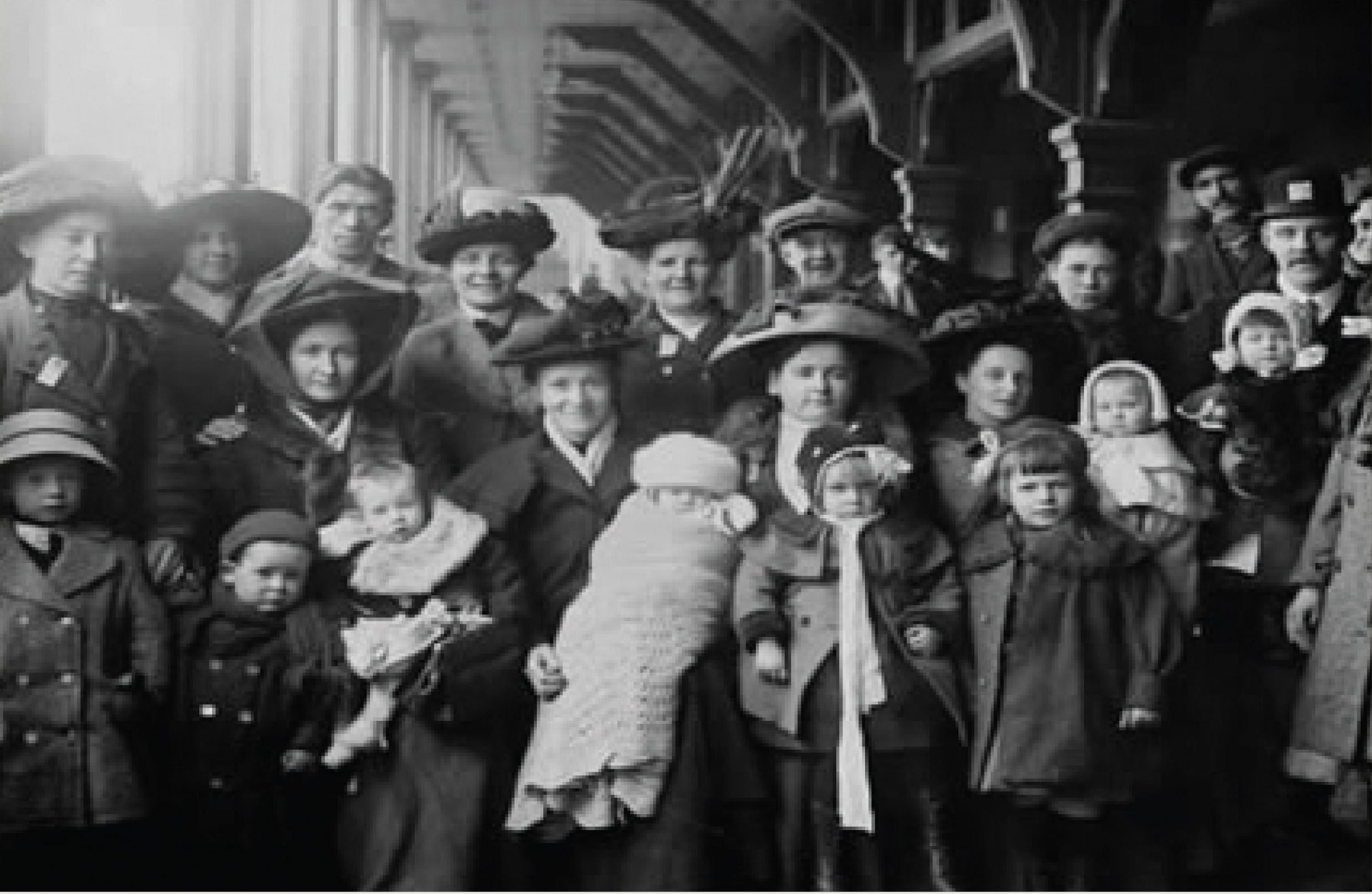 A group of immigrants poses for a photo at a train station while they wait for the train. Most are in winter coats and hats. Children are bundled in scarves and blankets.