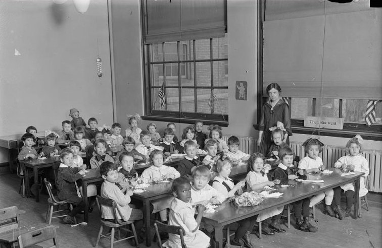 A large group of young children sit at rows of desks in a classroom and a young, female teacher is standing nearby.