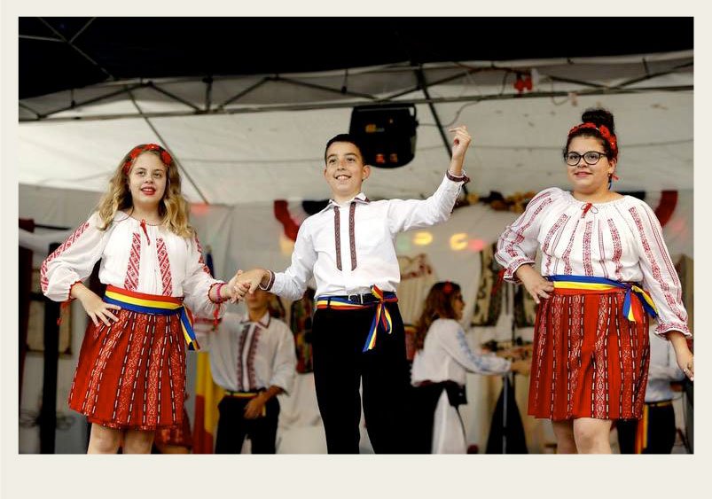 Past-Immigration-Romanian-dancetrio Two women and a man are dressed up in traditional Romanian outfits and are dancing on a stage.