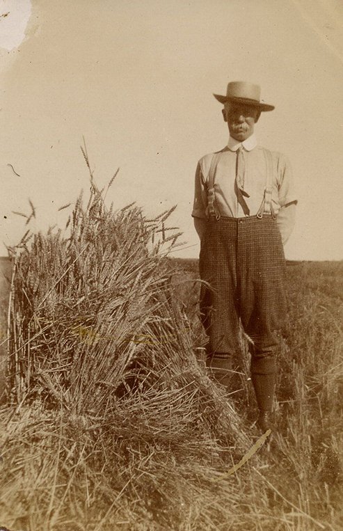 A farmer wearing a hat and a tie is standing next to a stook of wheat in a field.