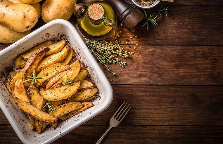 A casserole dish full of baked potatoes is on a table with other cooking ingredients including cooking oil, pepper, herbs, and potatoes.