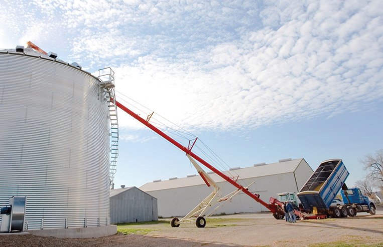 A grain truck unloads grain into a hopper, where the auger is pulling it from the truck to the top of a granary bin.