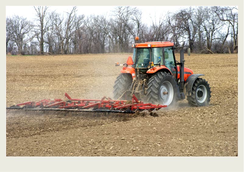 A tractor pulls a red cultivator in a field that has brown soil with no plants growing other than a grouping of trees in the background.