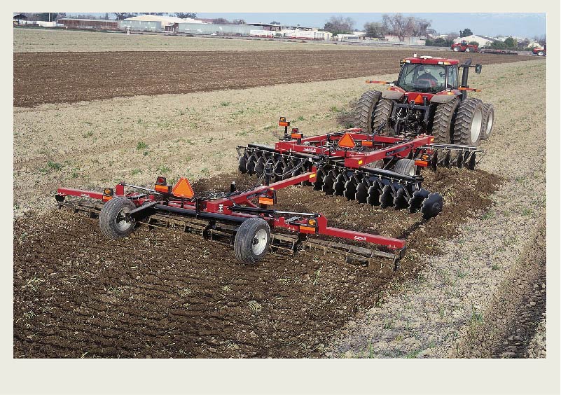A red tractor pulls a disker in a field next to a town that is in the background.
