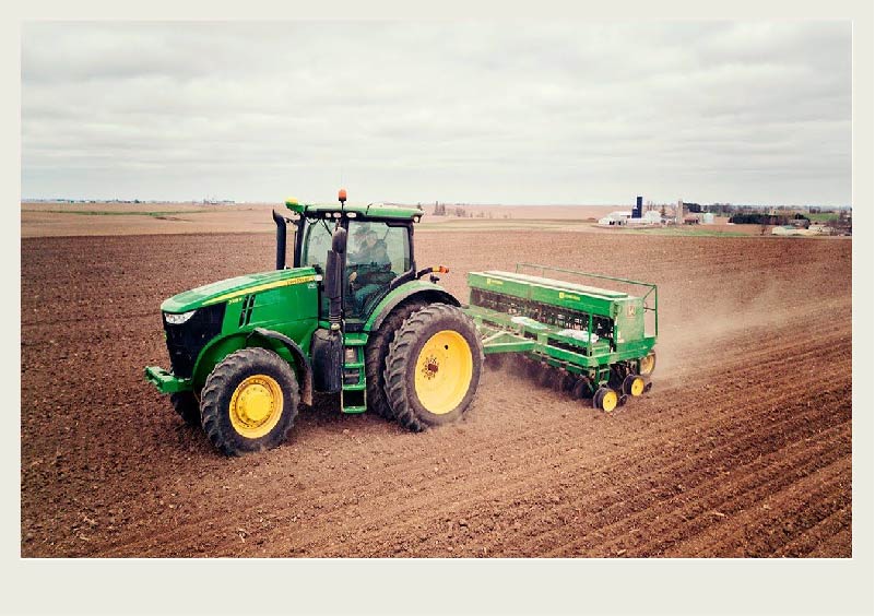 A tractor pulls a drill in a field with a farm yard in the background.
