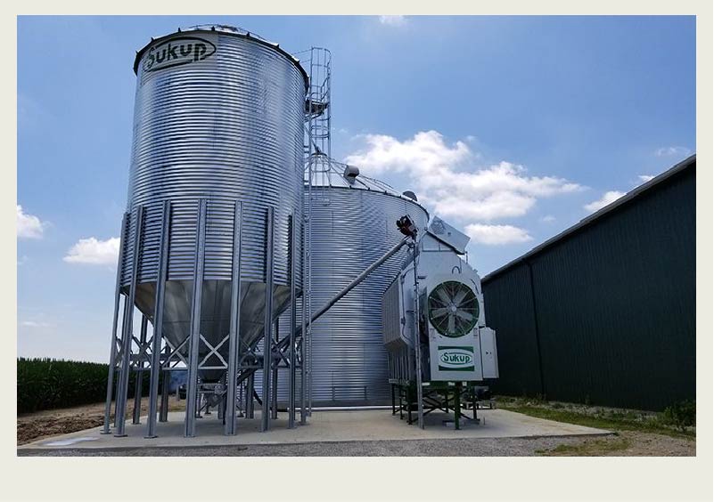 A grain dryer with a fan on the side of it is connected to the side of a grain bin. There are other grain bins in the background.