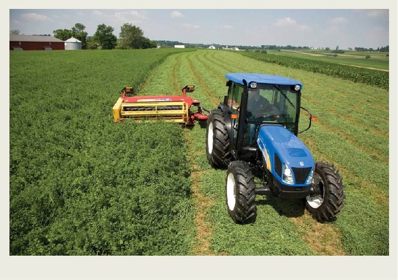 A blue tractor pulls a haybine in a field of hay. There are rows of hay that have not been cut by the haybine yet.