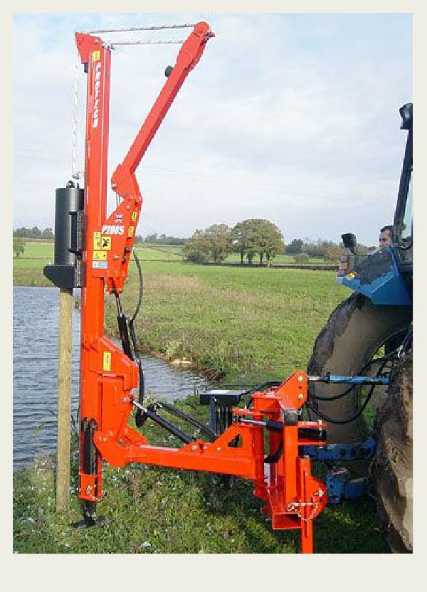 A post pounder is hooked up to a tractor to power it. The post pounder is being used to drive a post into the ground next to a dugout, out in a farm field.