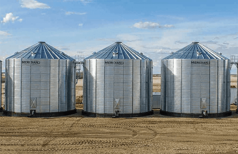 Three steel grain bins stand next to each other with a farm field in the background.