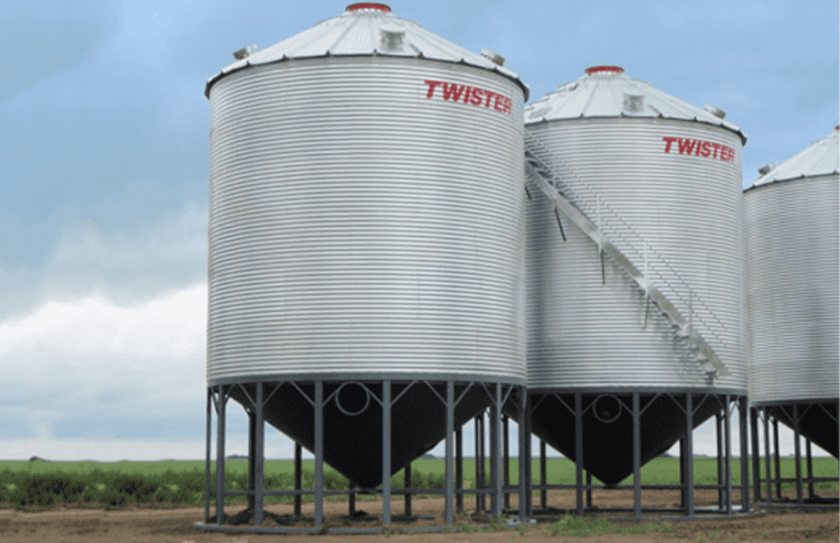 Three steel hopper bottom grain bins are next to a farm field.