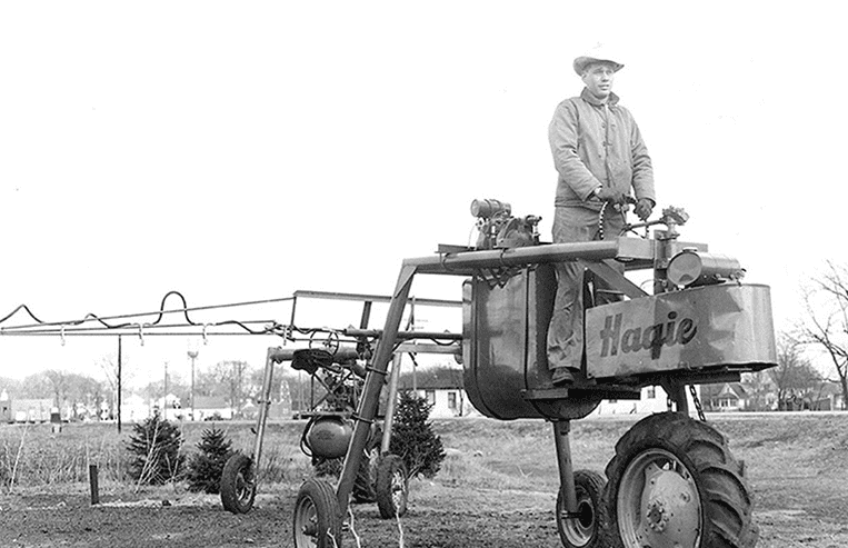 A farmer is standing on top of an open-cab high-clearance sprayer in a grassy area with a town in the background.