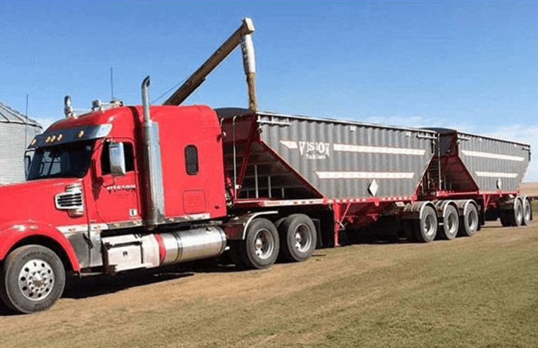 A semi-truck has a Super-B trailer attached to it which is getting loaded with grain that is dumping out of an auger.