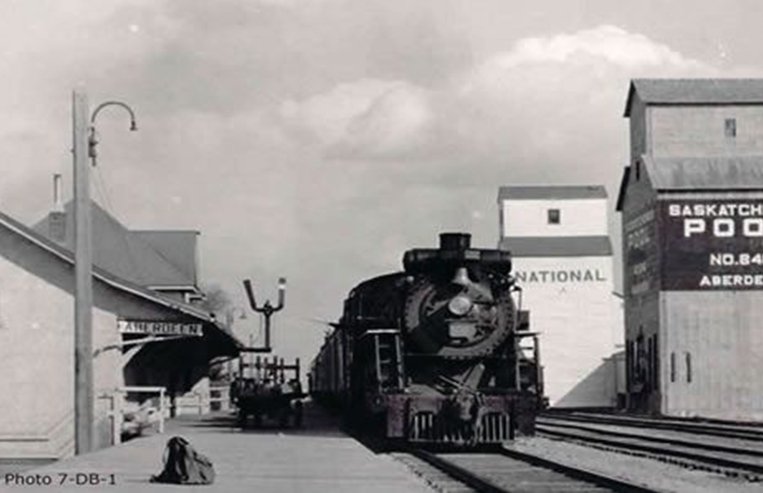 A train is stopped in a small town next to a train station and two elevators.