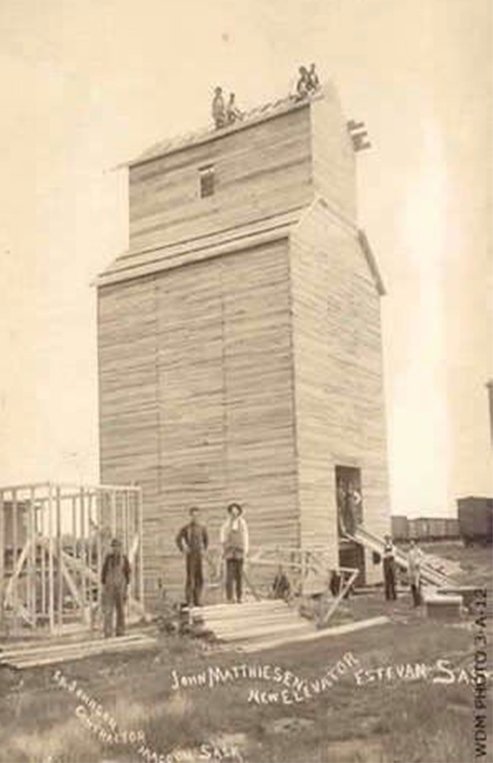 A group of men are working together to build a wooden grain elevator with piles of lumber nearby and a train in the background.