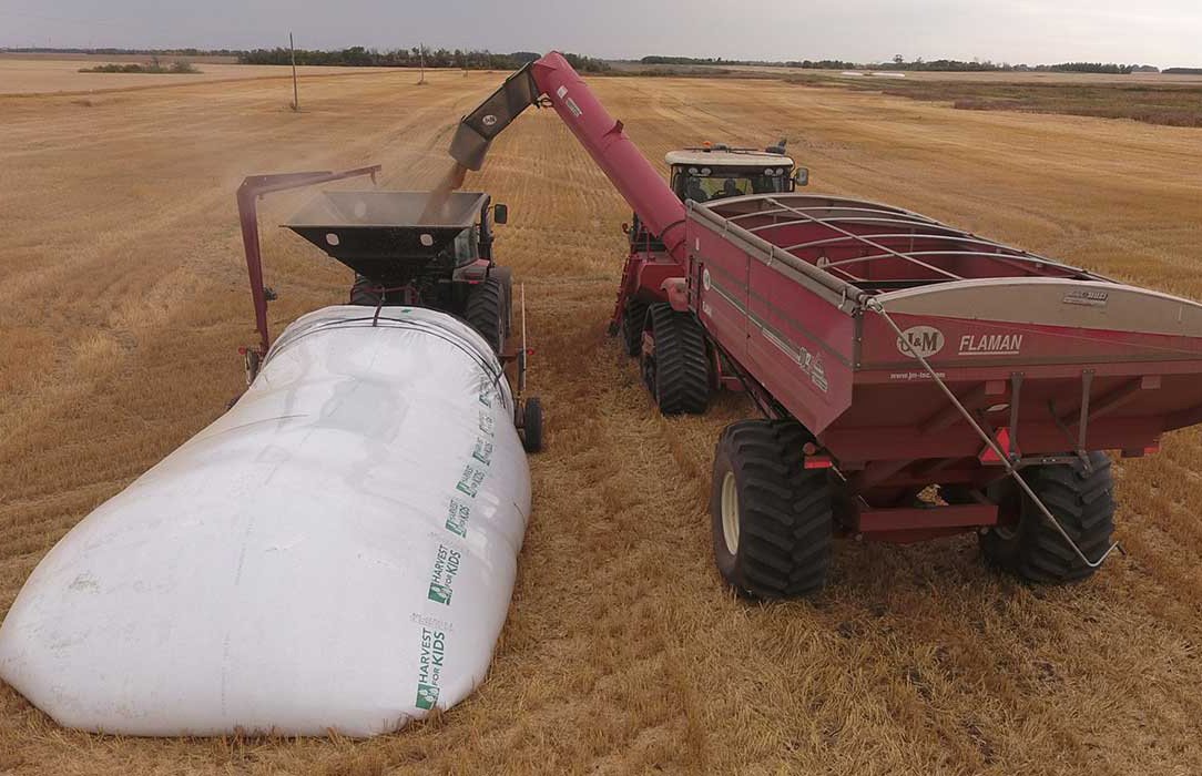 A large, white grain bag is in a field with a grain cart dumping grain into it.
